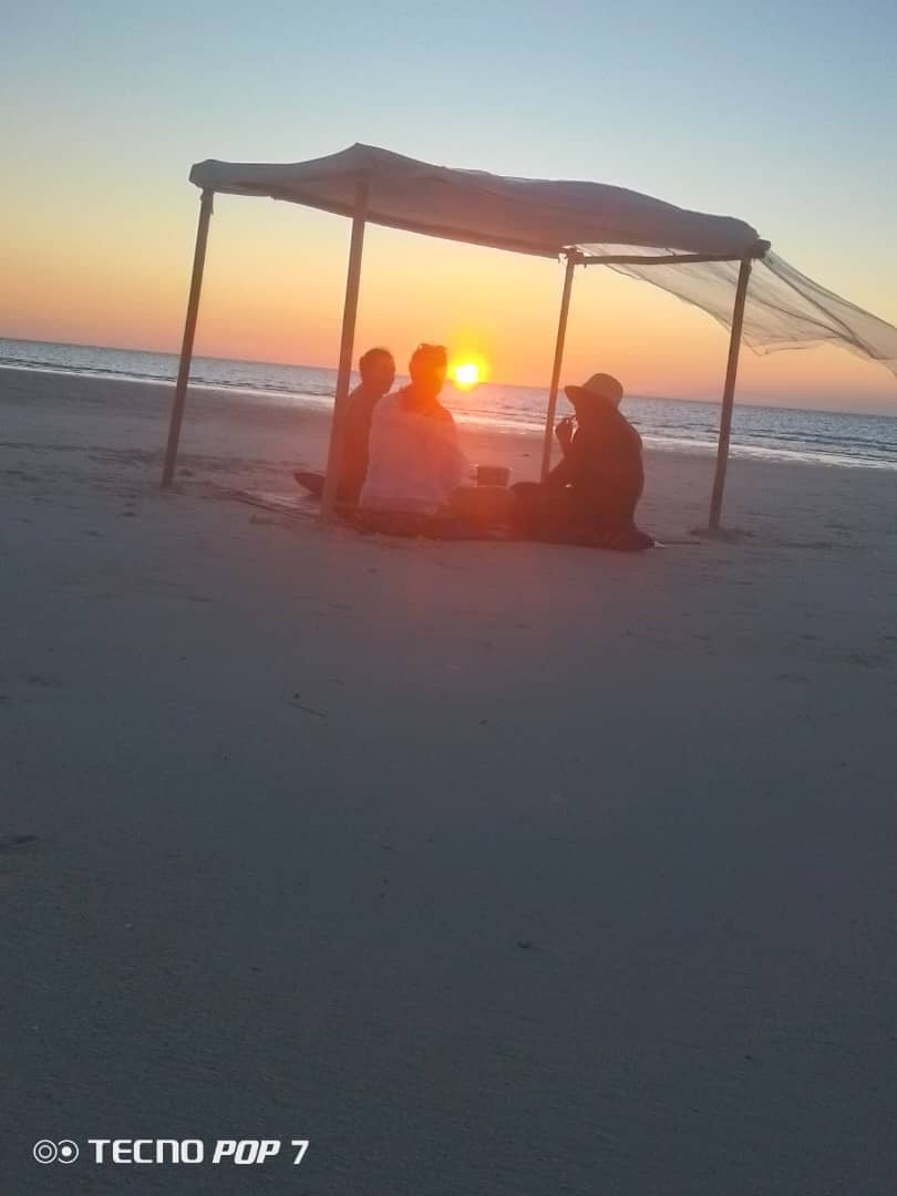 Two guests under a sandbank shade with the sun setting over the sea behind them