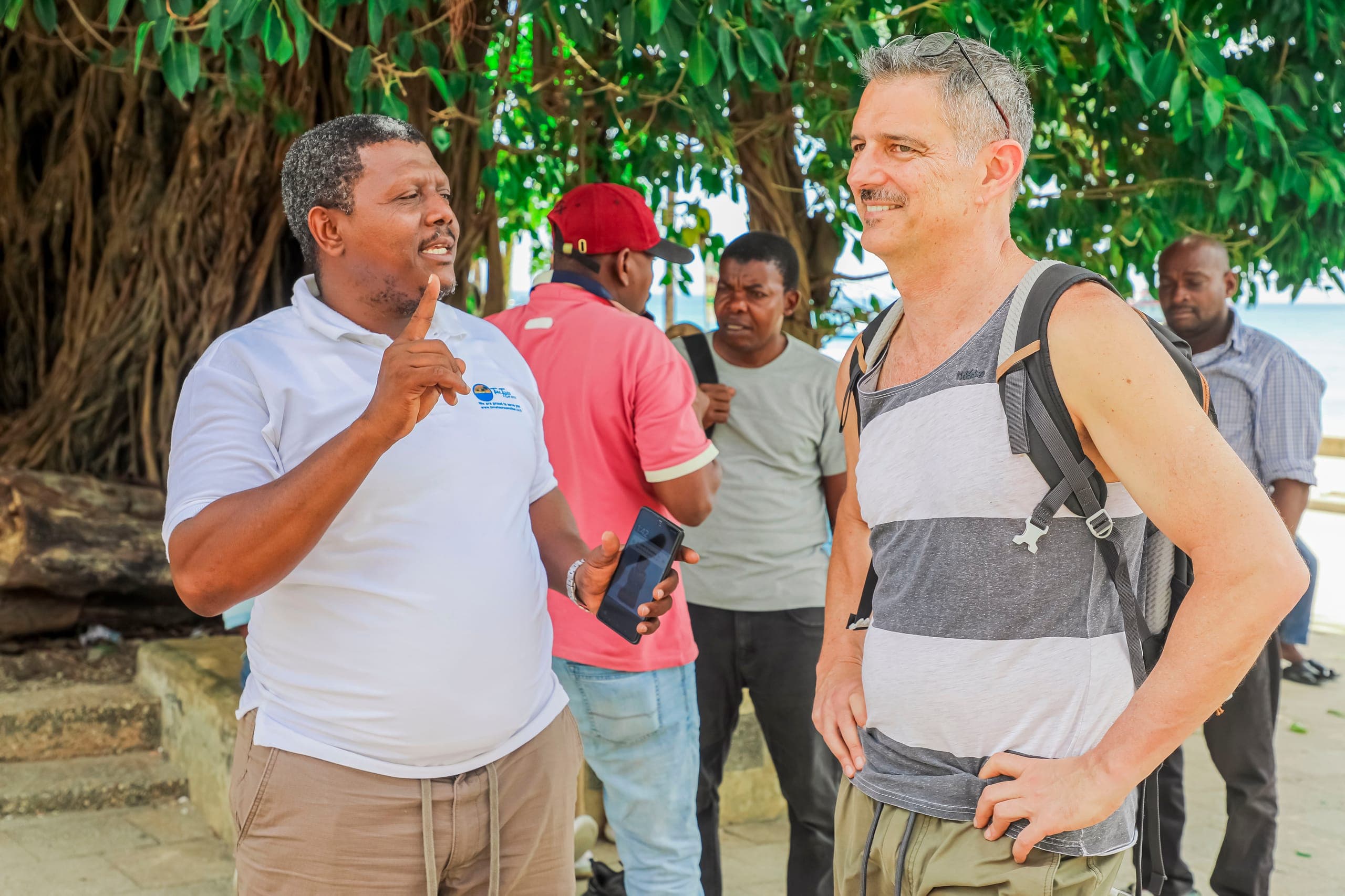 Salum, founder of Tima Tours, talking with a guest under a large tree on the Stone Town seafront