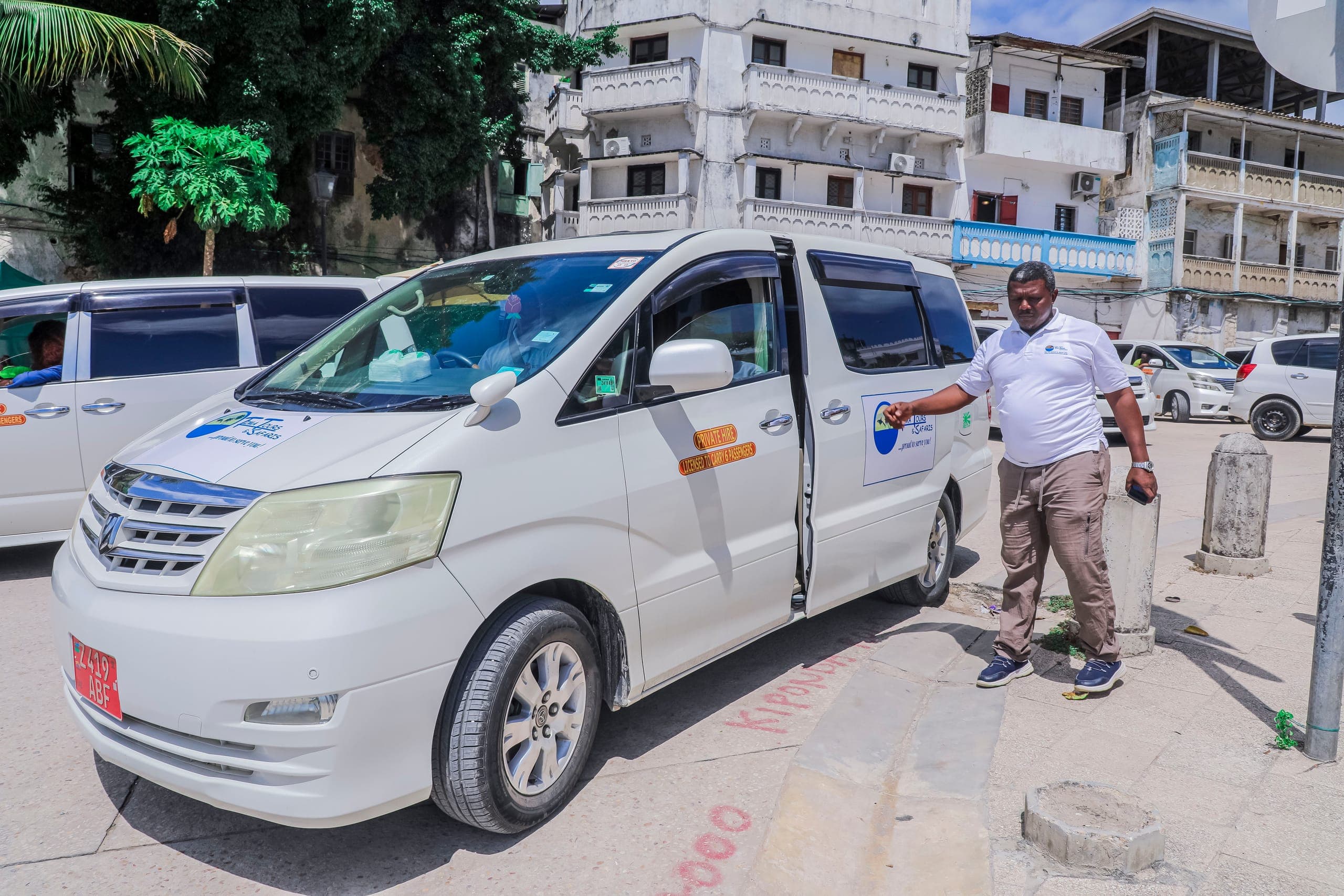 Salum beside a white Tima Tours minibus with its door open on a Stone Town street